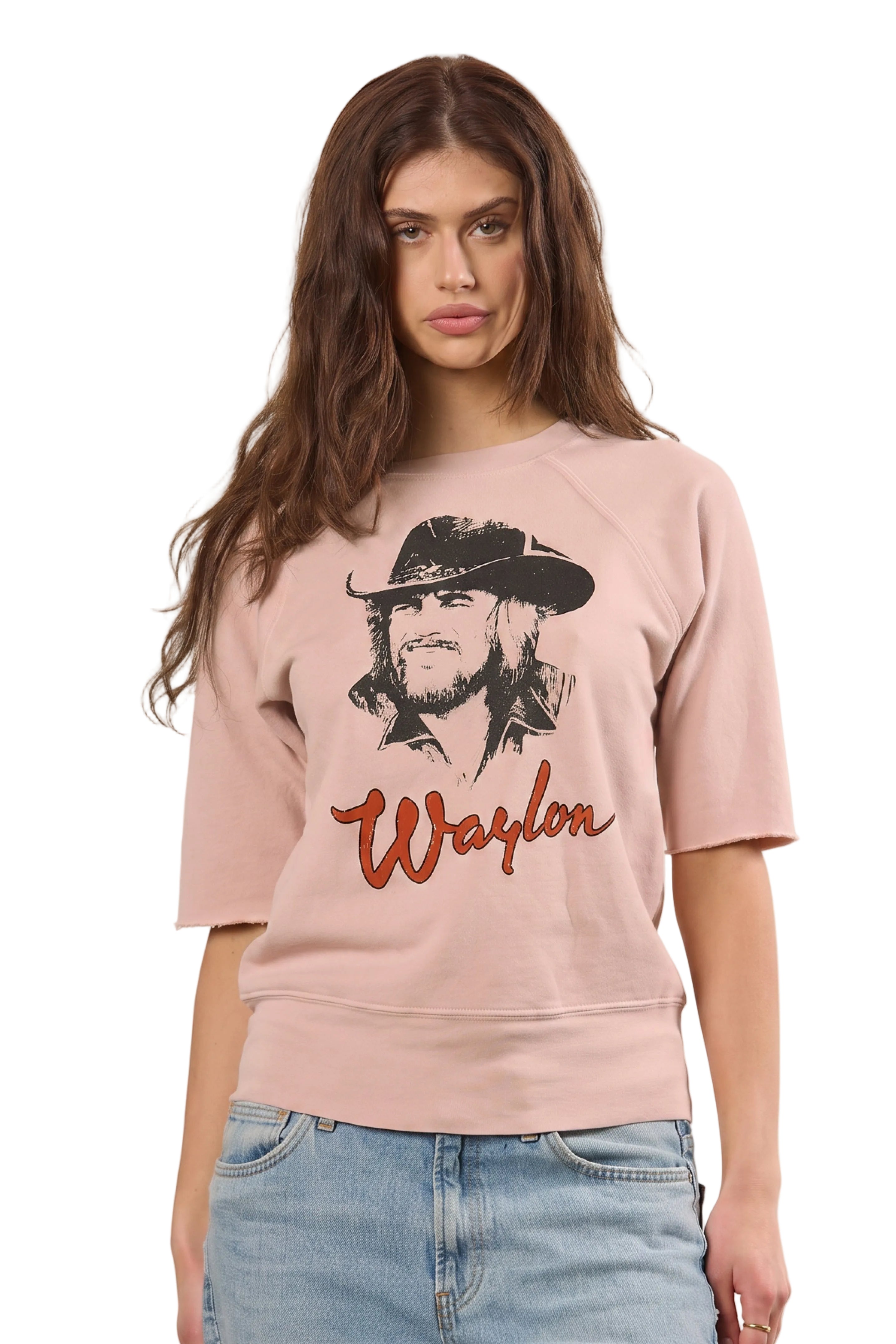 A woman modeling a light pink crewneck sweatshirt featuring a black graphic of Waylon Jennings and the name 'Waylon' in red script, paired with light blue jeans against a solid beige background.
