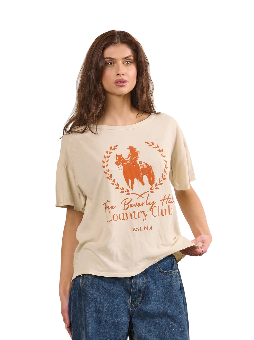 A woman modeling a beige short-sleeve graphic t-shirt with an orange print of a cowboy on a horse and the text 'The Beverly Hills Country Club', paired with blue jeans, against a solid beige background.