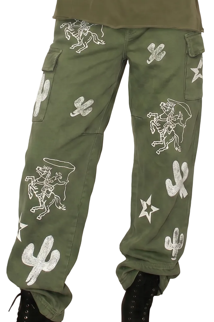 A model stands against a solid beige background wearing an olive green sleeveless top, matching olive green cargo pants with white desert-themed prints (cacti, stars, skulls), and black lace-up boots.