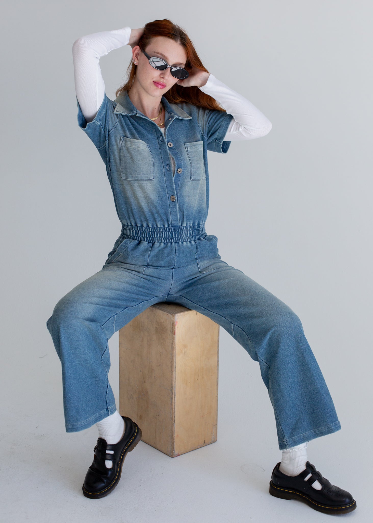 Person wearing a blue denim jumpsuit sitting on a wooden block against a plain background